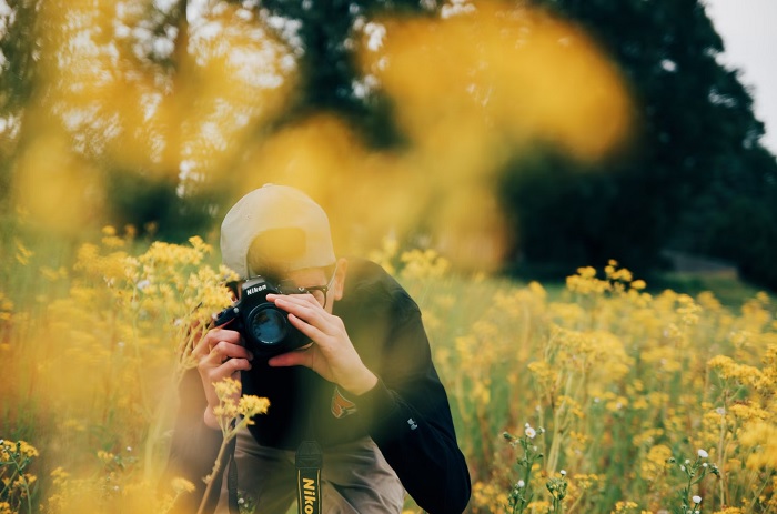 Young man with a camera taking a photo in a meadow of yellow flowers