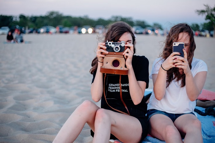 Two girls photographing with two of the best cameras for teens while sitting in a beach