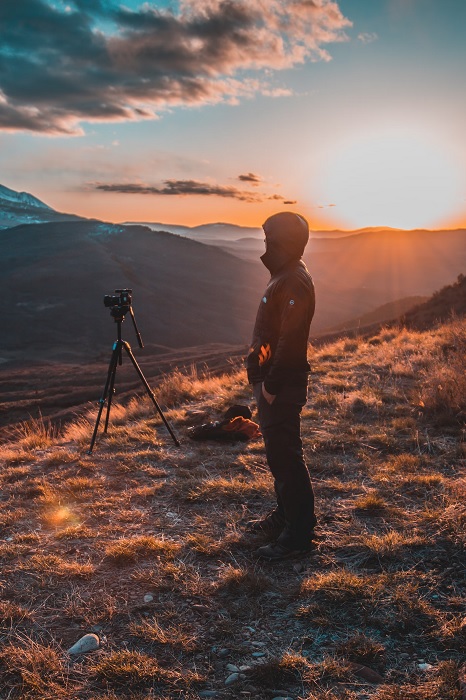 Man standing near a camera on a tripod on a mountain side at sunset