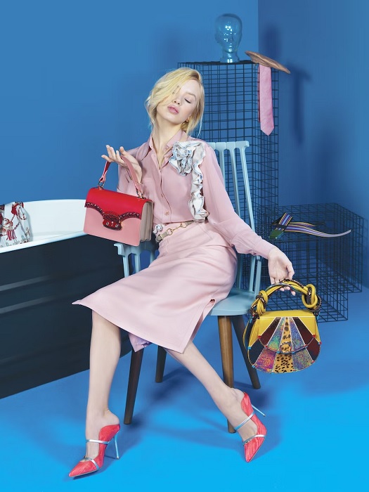 Woman in a pink skirt-suit holding two handbags in a blue room