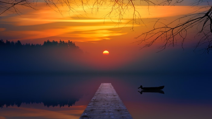Sun setting over a foggy lake with trees, a dock, and a boat