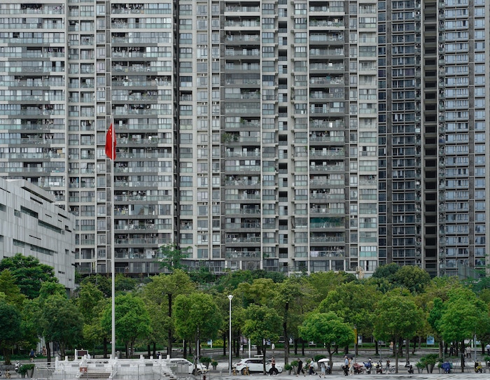 Cityscape with tall, gray city apartment buildings in the background and green trees and tiny cars and people in the foreground