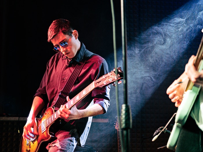 A guitarist wearing sunglasses and playing an electric guitar with a spotlight in the background