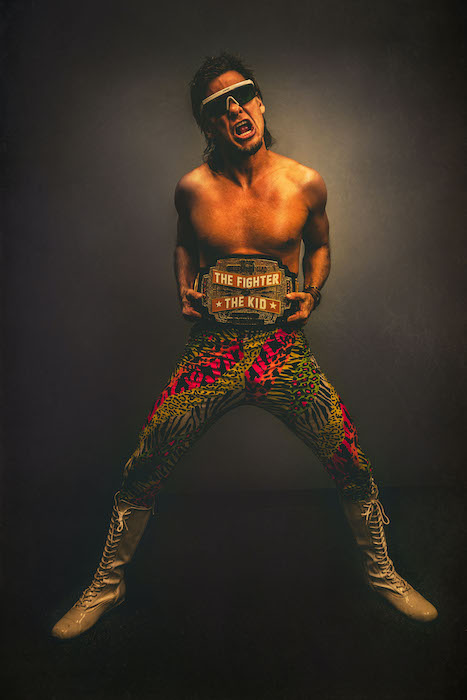 A wrestler posing with sunglasses, colorful pants, and a wrestling belt