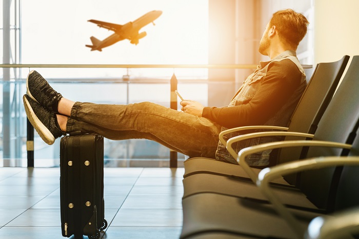 A man sitting with his feet up on a suitcase in an airport watching a plane fly by